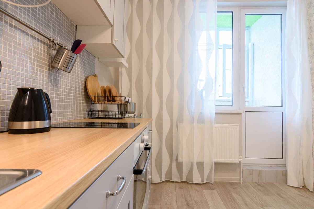 A narrow kitchen area with wooden countertop, wooden flooring and whit patterned curtains
