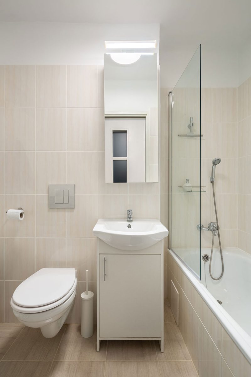 A narrow bathroom with white vanity and a glass wall and bathtub