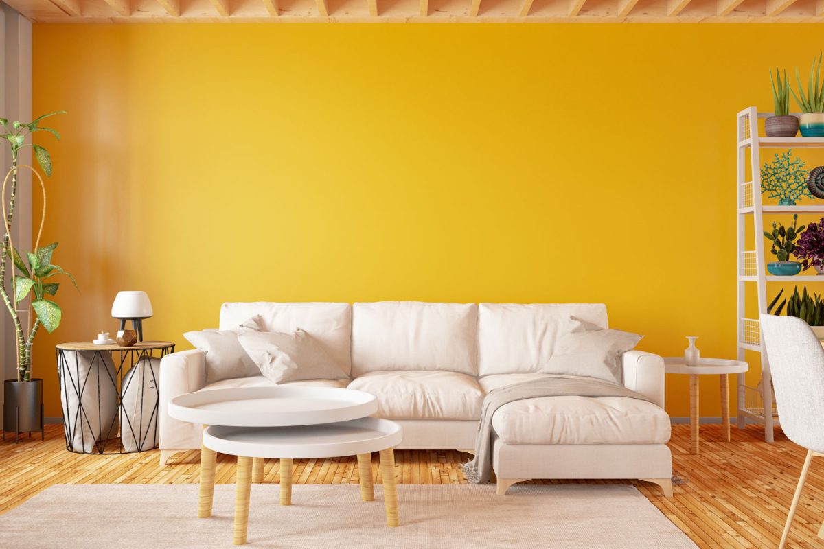 A yellow wall with a white sectional sofa with white throw pillows and a plants for liveliness