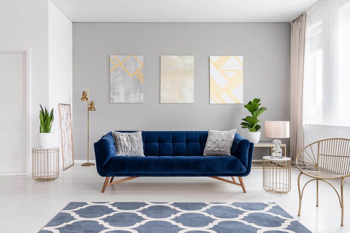 An elegant navy blue sofa in the middle of a bright living room interior with gold metal side tables and three paintings on a gray wall