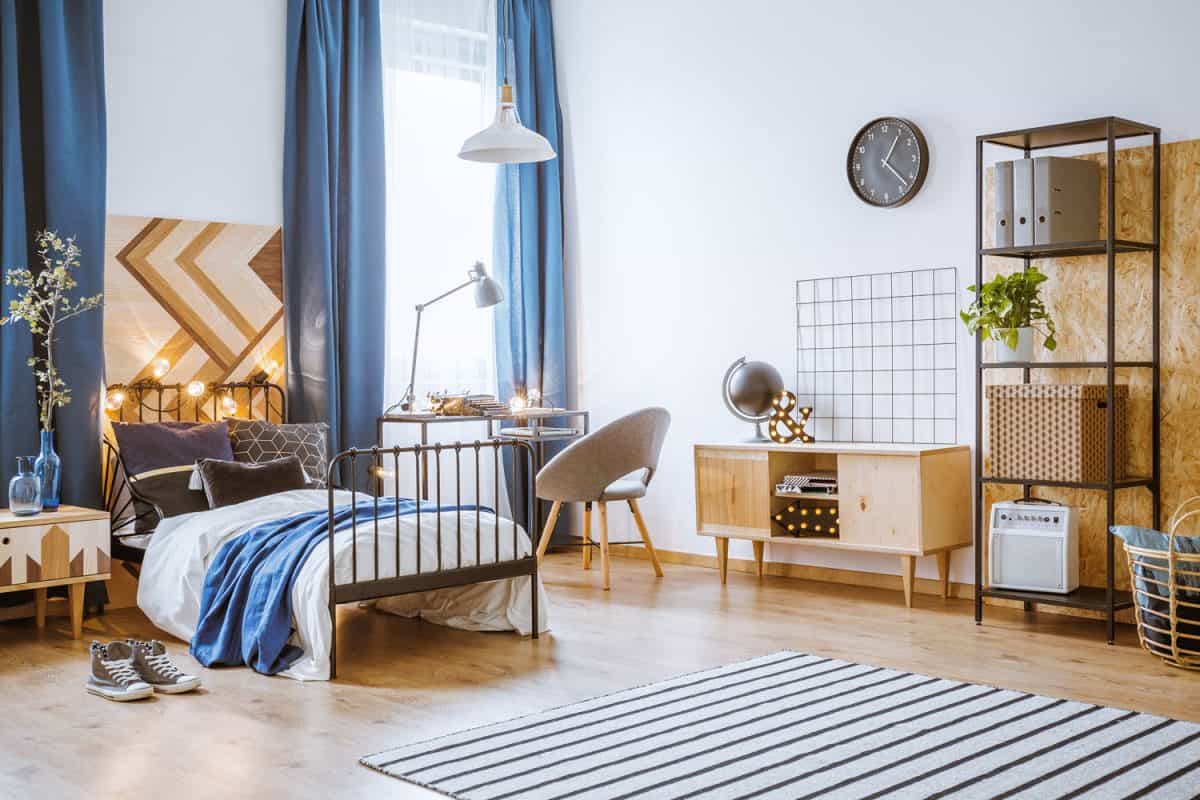 Bright bedroom interior for a boy with an iron bed, wooden cupboard, rug, light decorations, black clock and navy blue curtains