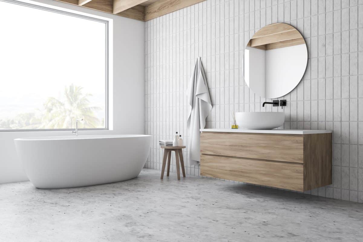 Corner of hotel bathroom with white and tiled walls, concrete floor, comfortable bathtub standing under window and sink on wooden countertop with round mirror.