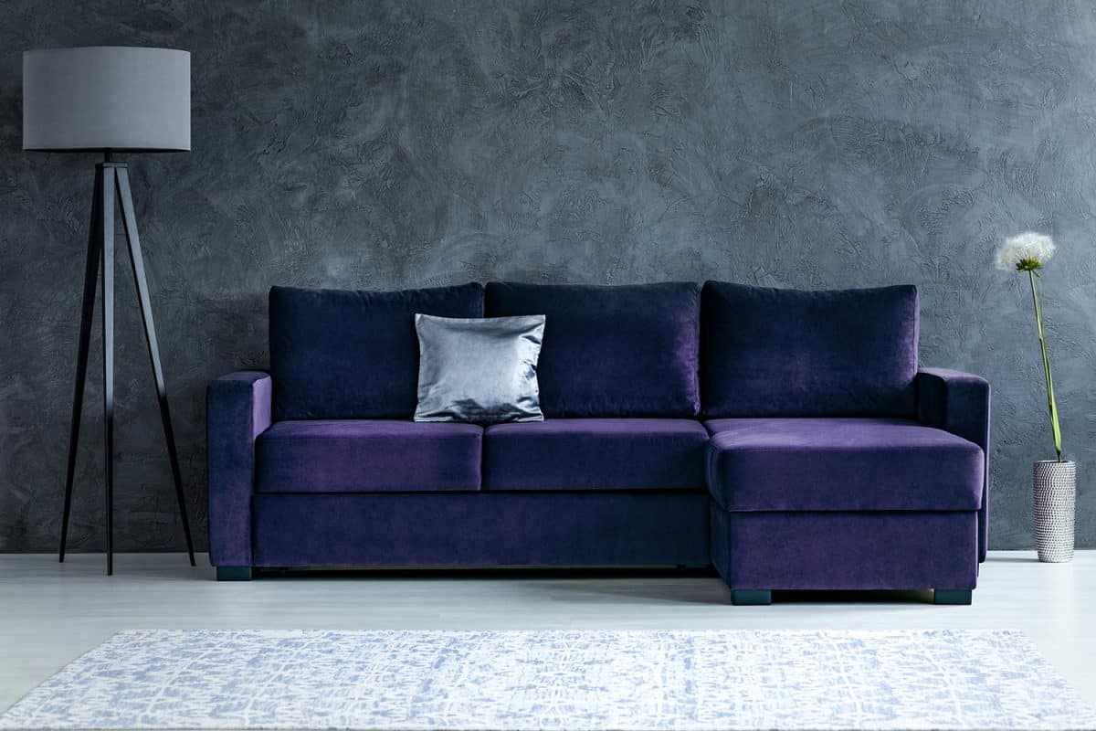 Dandelion next to purple corner sofa with silver pillow in dark living room interior with lamp