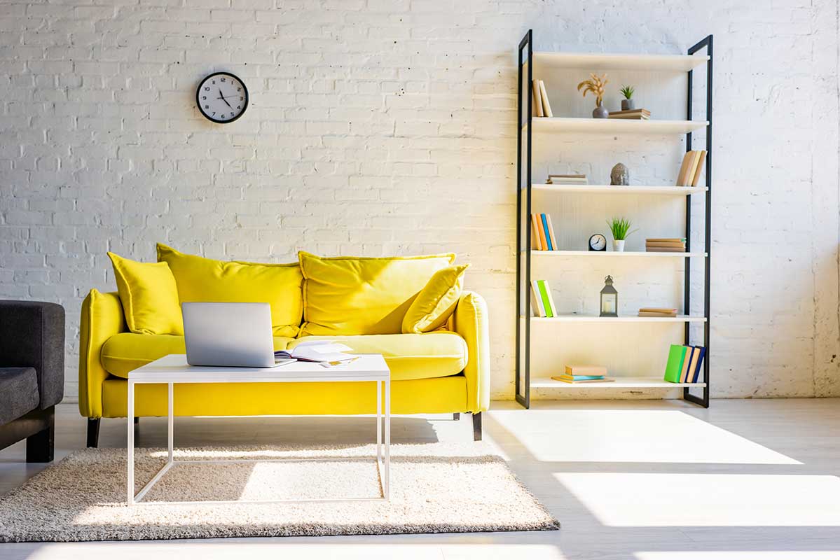 Living room with yellow sofa, shelf and table with laptop in sunlight