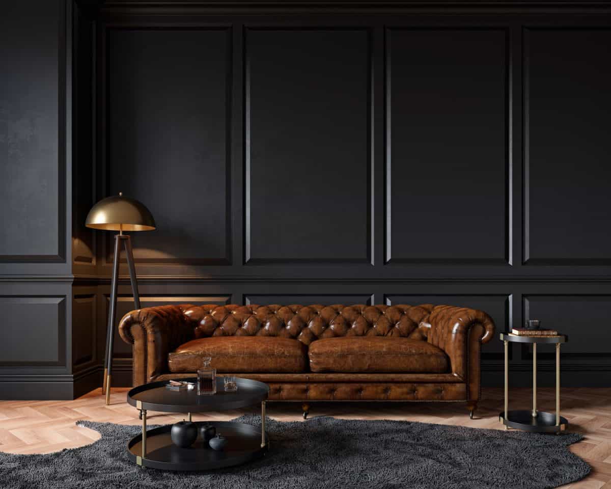 Modern classic black interior with capitone brown leather chester sofa, floor lamp, coffee table, carpet, wood floor, mouldings. 