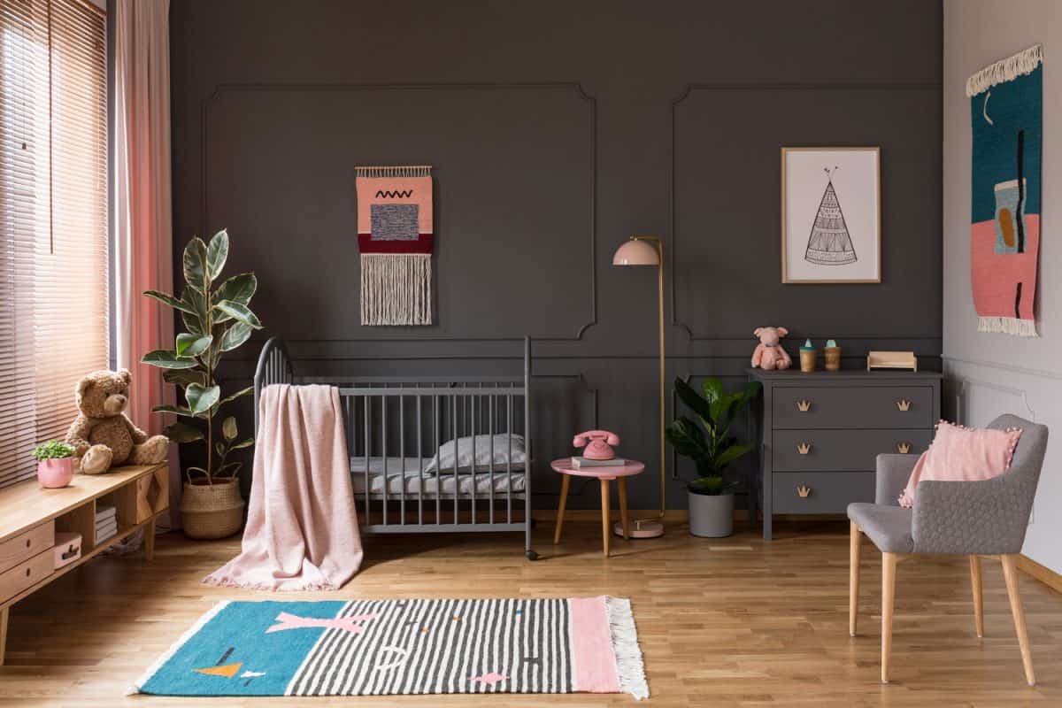 Real photo of a grey crib standing next to a pink stool, a lamp and cupboard in grey baby room interior also with armchair, rug and posters
