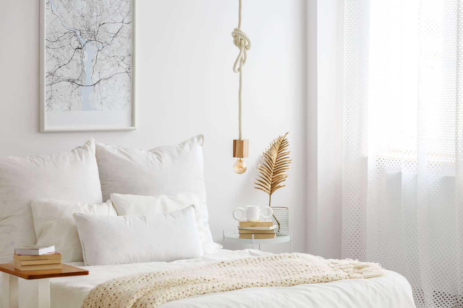 Simple bulb lamp on a rope hanging above bed with white bedclothes, books and gold fern leaf on an end table in white bedroom interior, What Curtains Go With White Walls?
