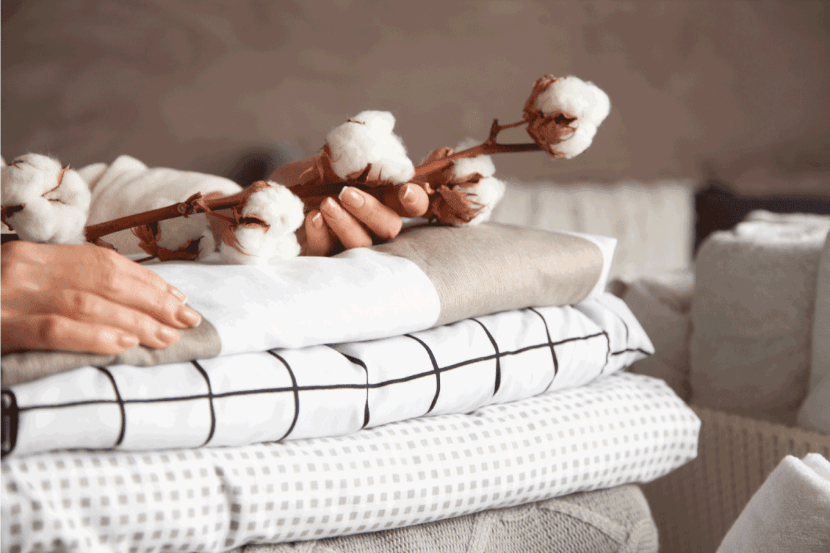 Well groomed woman hands holding the cotton branch with pile of neatly folded bed sheets, blankets and towels.