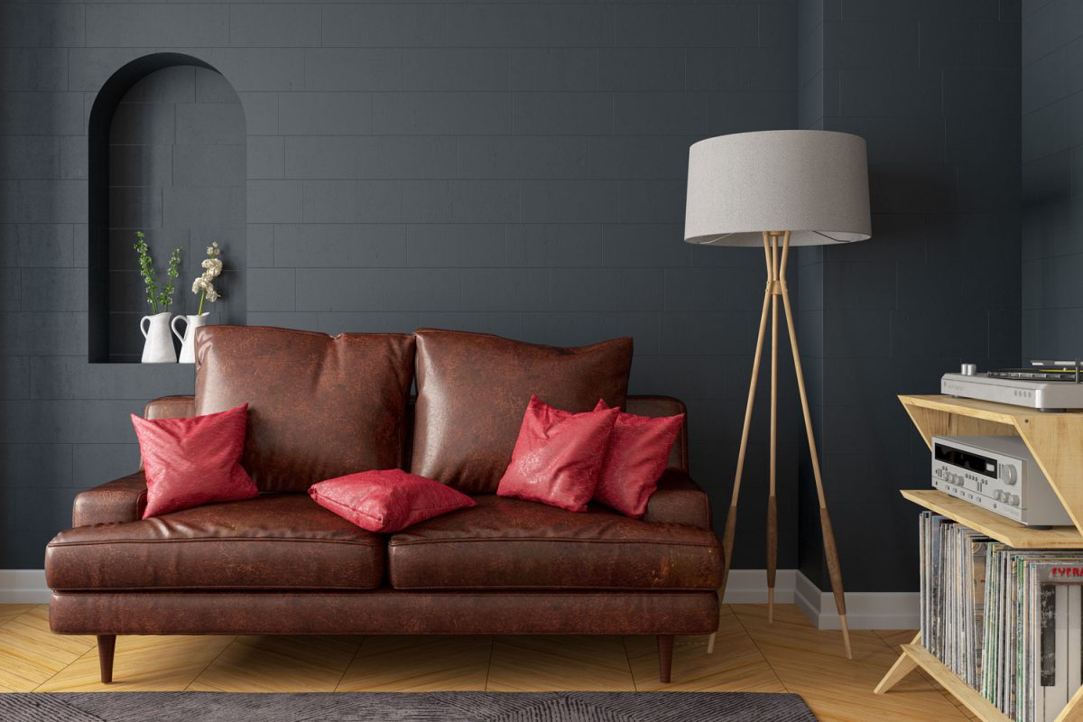 A brown leather sofa with red throw pillows and a black wall with a tripod lamp