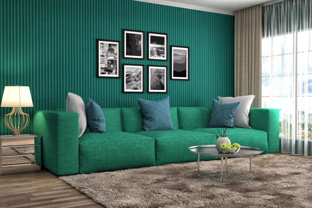 A long green sofa with light blue and white throw pillows a carpet on the front matched with a green wall paper on the wall with paintings