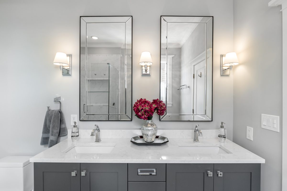 A luxurious bathroom with a grey double vanity with flowers sitting on the white quartz counter top.