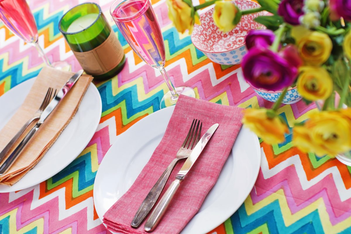 Bright colorful table setting with multi colored chevron pattern tablelcoth, pink champagne and colourful napkins. Roses in pink and yellow blurred in foreground and green scented candle decoration

