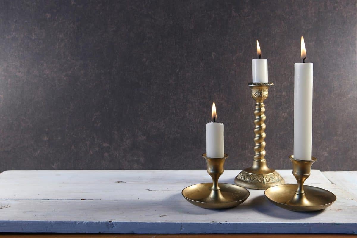 Burning candles in vintage metal candlesticks on white wooden table against dark stone background