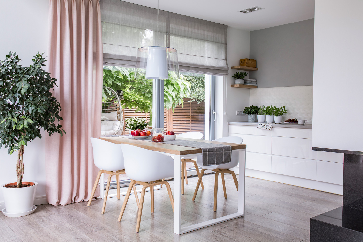 Gray roman shades and a pink curtain on big, glass windows in a modern kitchen and dining room interior with a wooden table and white chairs.