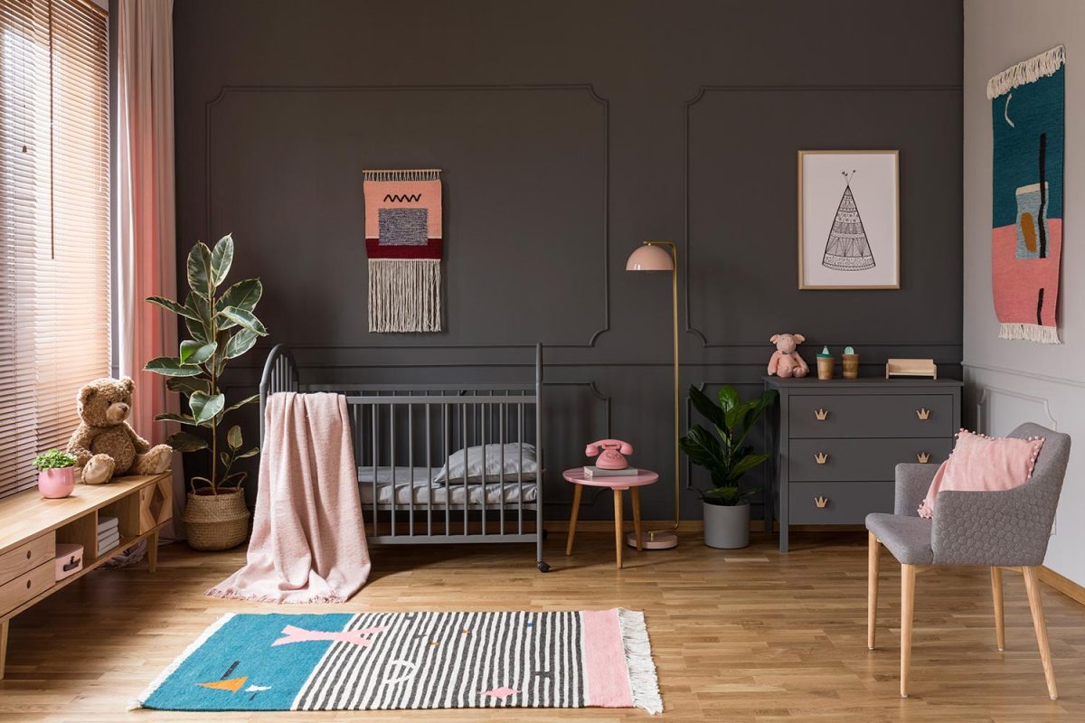 Grey crib standing next to a pink stool, a lamp and cupboard in grey baby room