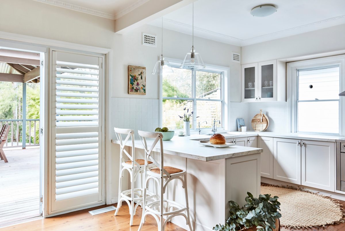 Interior photography of a fresh white Hamptons style kitchen with breakfast bar, cross back bar stools, polished floor boards and a door way opening up to a back deck