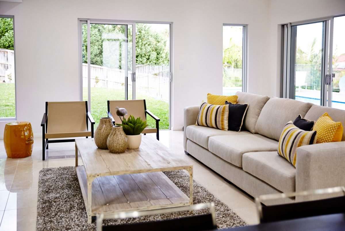 Interior photography of a modern lounge room with a beige sofa, cushions, arm chairs and coffee table with a rug, decorative objects and a garden in the background