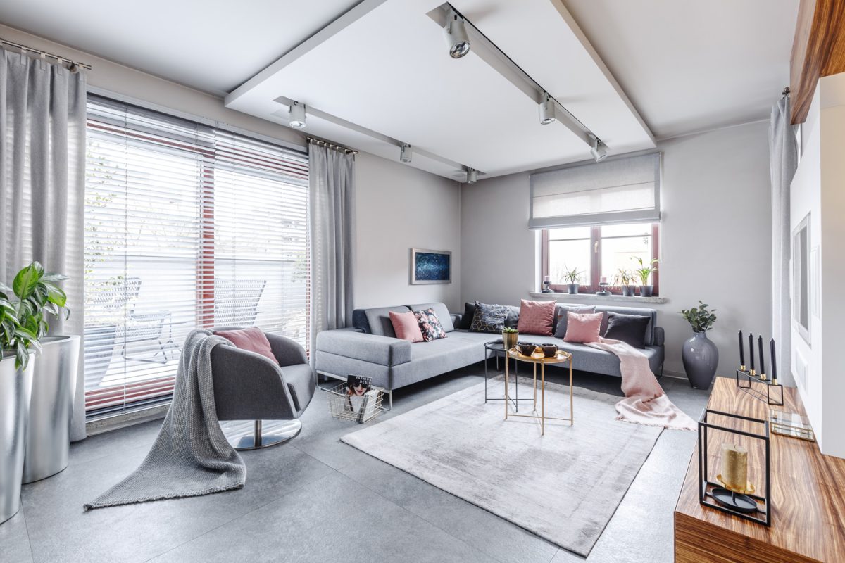 Modern interior with large gray corner sofa and armchair decorated with pillows and blankets in a bright cosy living movie room with light from big glass door to balcony, wooden elements and decorations

