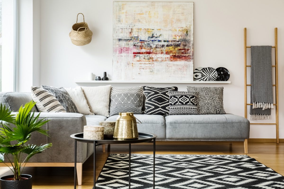 Patterned pillows on grey corner sofa in living room interior with table and painting.
