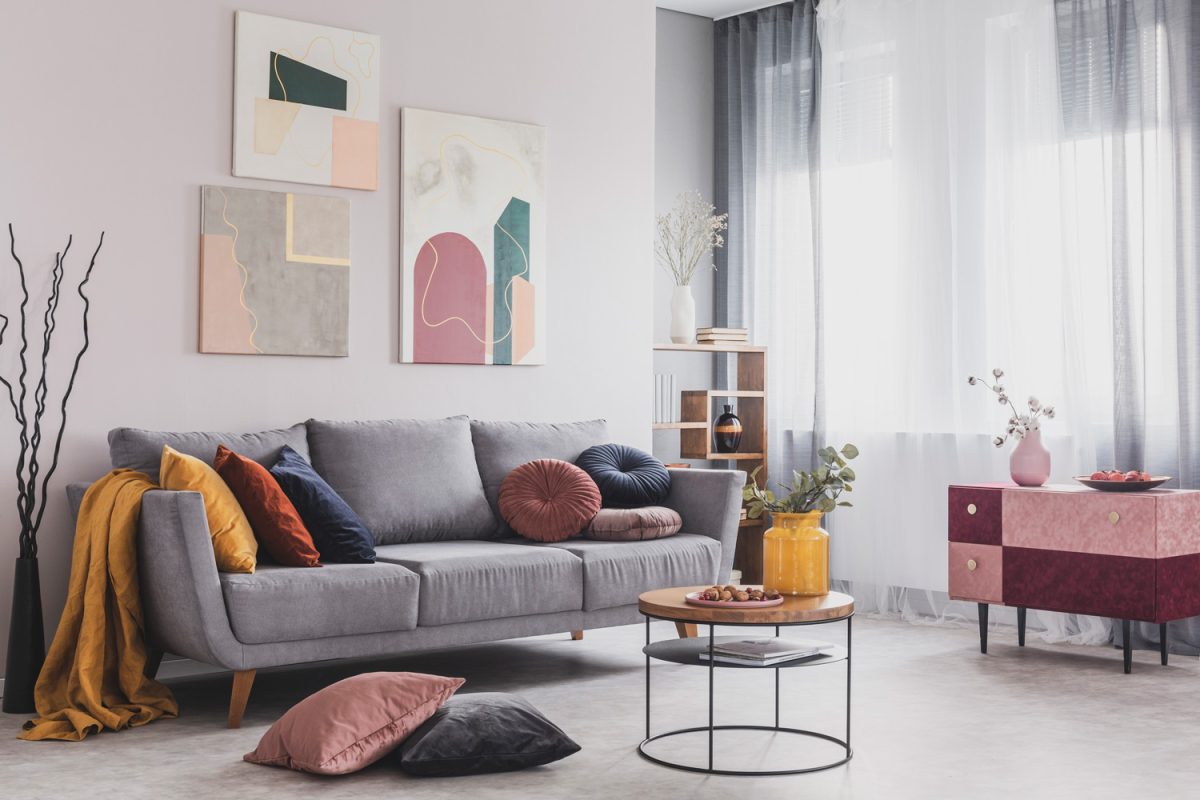 Real photo of abstract paintings hanging on white wall above a gray sofa in a living room interior with big windows

